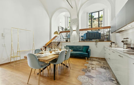 Modern apartment interior with a wooden dining table, blue chairs, teal sofa, geometric-patterned floor tiles, and arched windows.