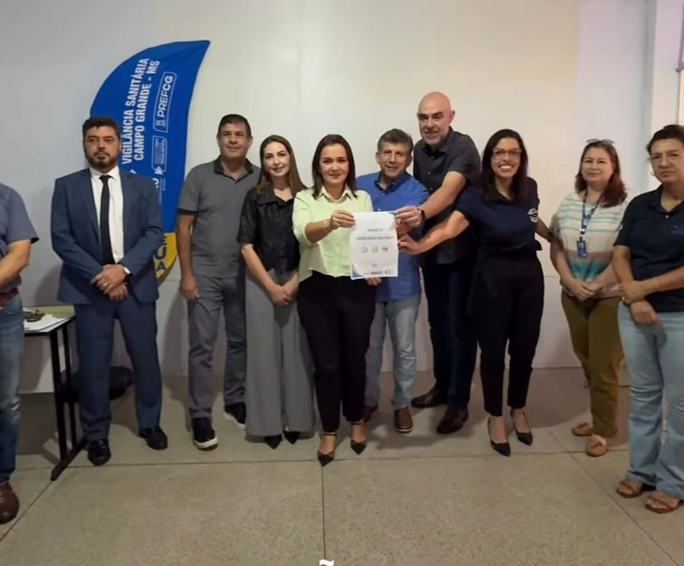 Grupo de pessoas ( vereador Beto Avelar, do Secretário Municipal de Saúde, Dr. Marcelo Luiz Brandão Vilela, além de representantes do DECON, PROCON, integrantes da Associação do Mercadão Municipal) sorriem para a câmera, segurando um documento (assinatura do projeto mercadão em foco). Banner azul diz "Vigilância Sanitária". Ambiente interno com piso claro.