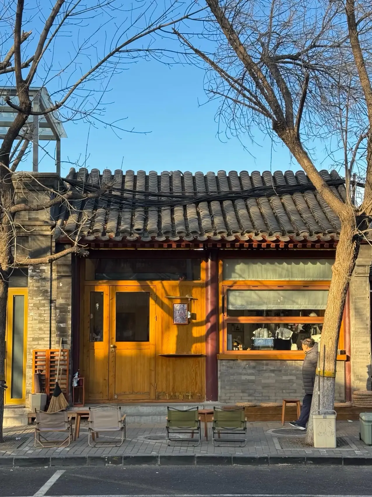 Traditional Beijing hutong street scene with grey brick courtyard house, wooden storefront, tiled roof, and small outdoor seating along the sidewalk.