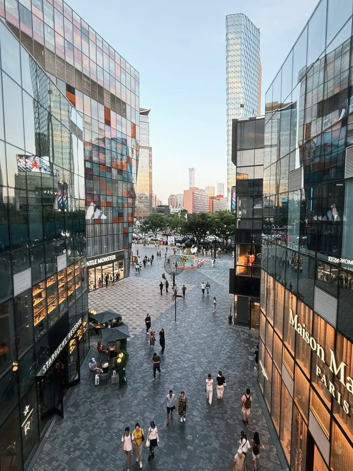 Open-air shopping street at Sanlitun Taikoo Li in Beijing with people walking between modern glass buildings, representing trendy Beijing shopping and lifestyle scenes.