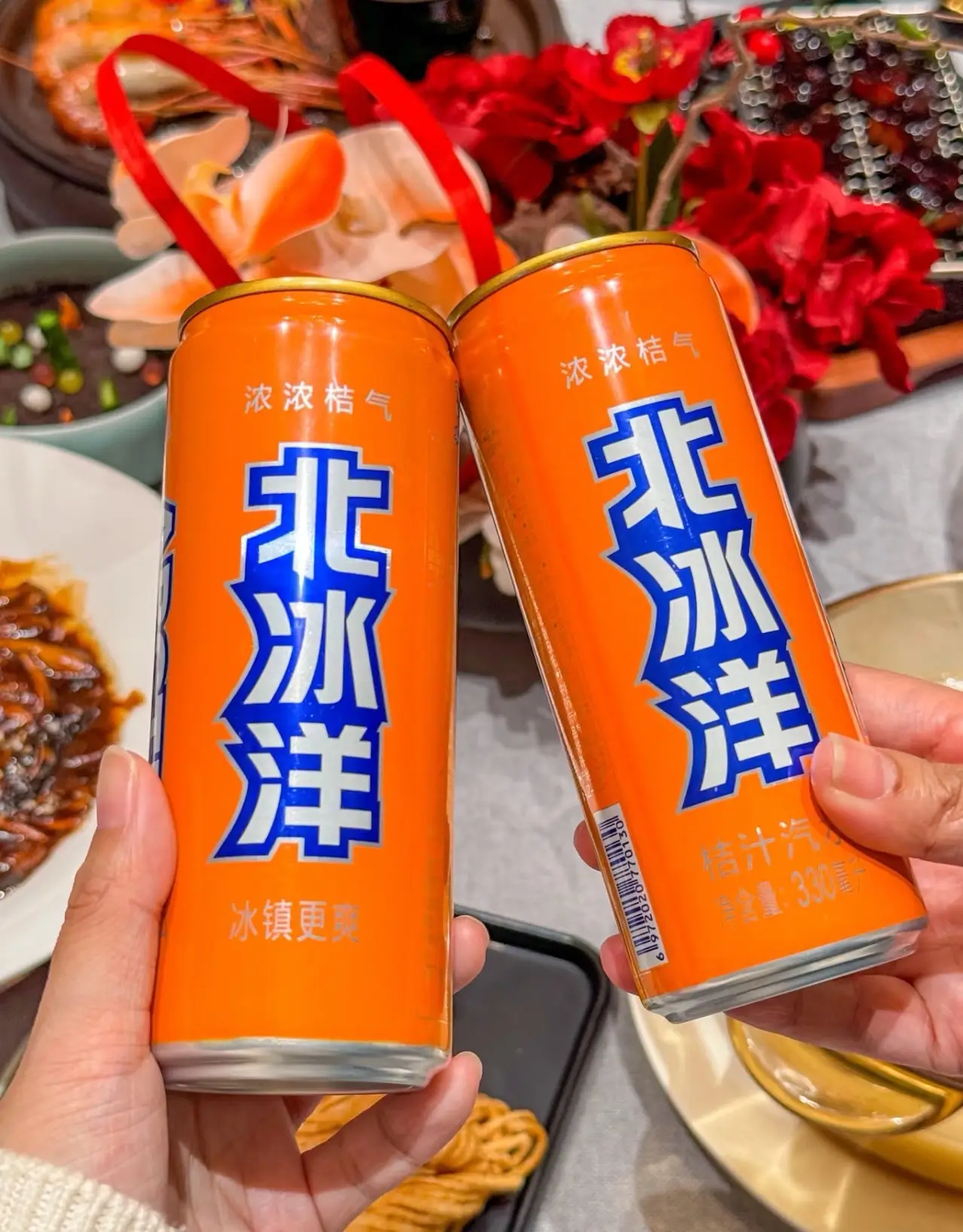 Two hands holding tall, bright orange cans of "Beibingyang" (Arctic Ocean) orange soda at a dining table. The classic Chinese soda brand features bold blue and white characters.