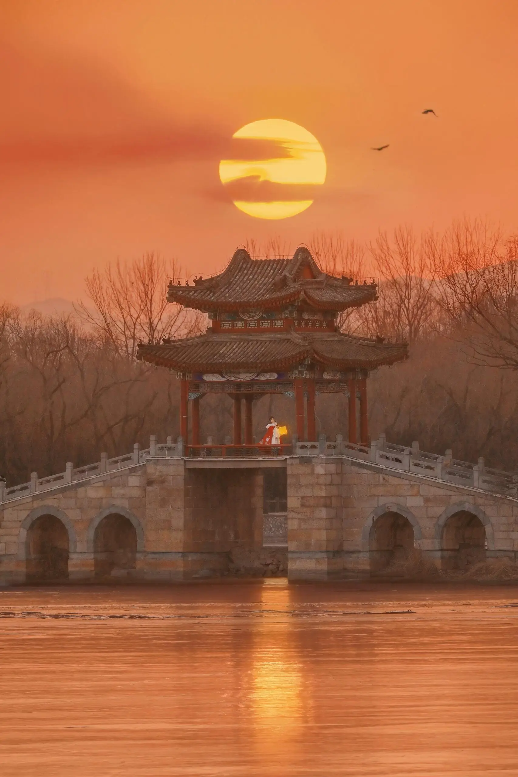 Historic pavilion standing on an arched stone bridge over calm water at sunset, framed by bare trees with the sun glowing in the background