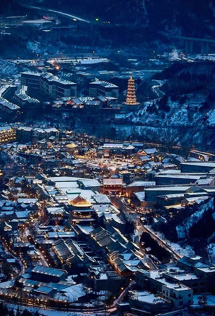 Aerial winter view of Gubei Water Town covered in snow, traditional rooftops and a lit pagoda surrounded by mountains at dusk near the Great Wall.