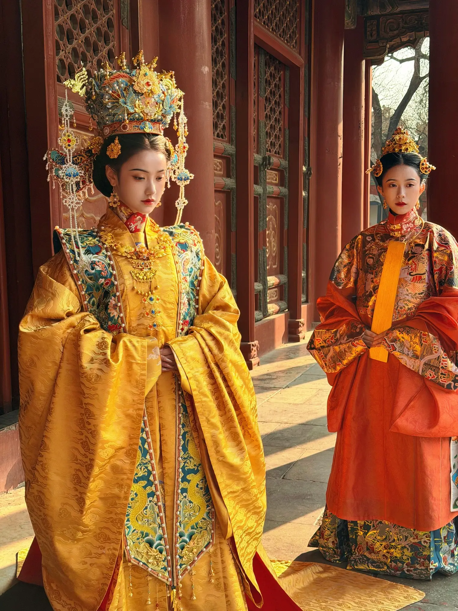 Two people wearing ornate imperial-style robes and elaborate headdresses stand in a palace courtyard, showcasing traditional Chinese royal attire against historic architecture.