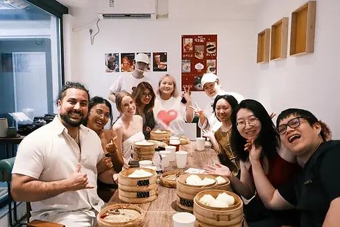A small group of travelers smiles around a table during a Beijing hutong bun-making experience, with bamboo steamers filled with freshly made baozi in front of them.