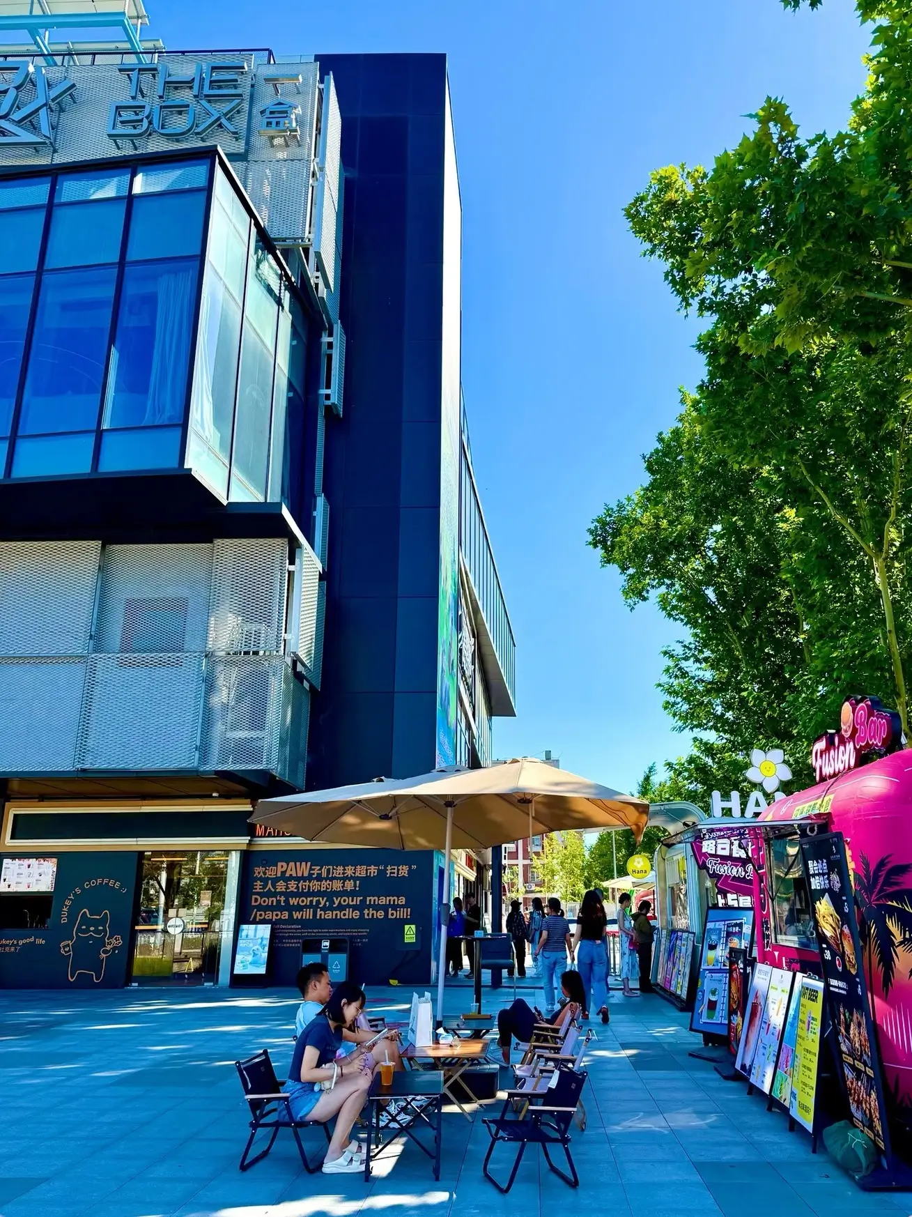Outdoor street scene at THE BOX Youth Energy Center in Beijing, featuring modern industrial-style buildings, cafés, and colorful food trucks with people relaxing in a trendy open-air shopping area.