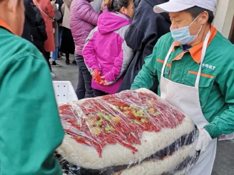Baiji staff moving a large traditional rice cake while customers wait outside the shop on Niujie Street