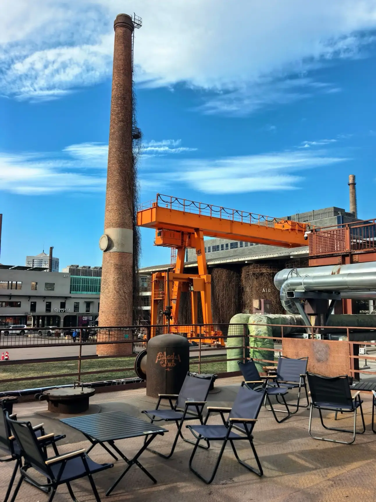 An open-air industrial terrace at the 798 Art District under a bright blue sky. The view features a tall brick factory chimney, a massive orange industrial crane, and exposed ventilation pipes. In the foreground, a modern seating area with black folding chairs and tables offers a contemporary contrast to the historic industrial ruins.