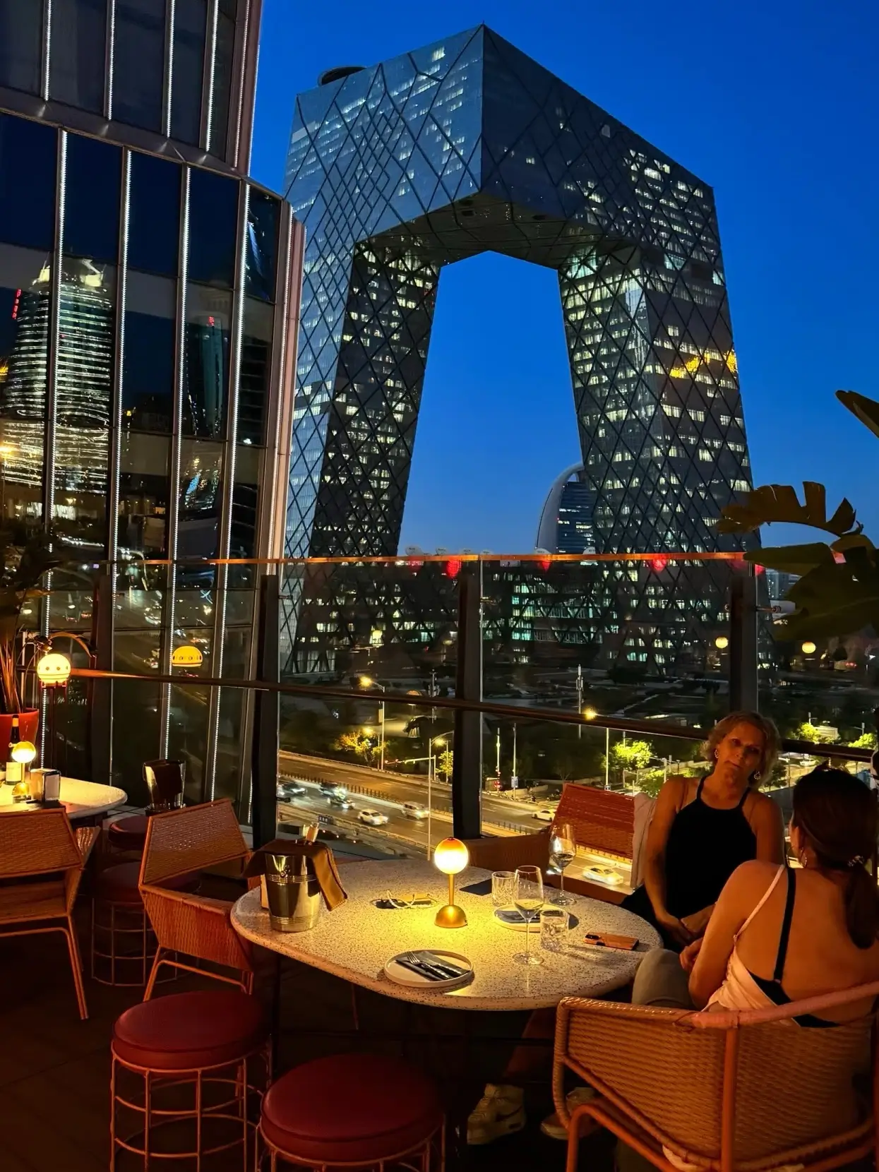 Night view of CCTV Headquarters from a rooftop restaurant in Beijing CBD, with city lights and diners enjoying skyline views near China World Mall.