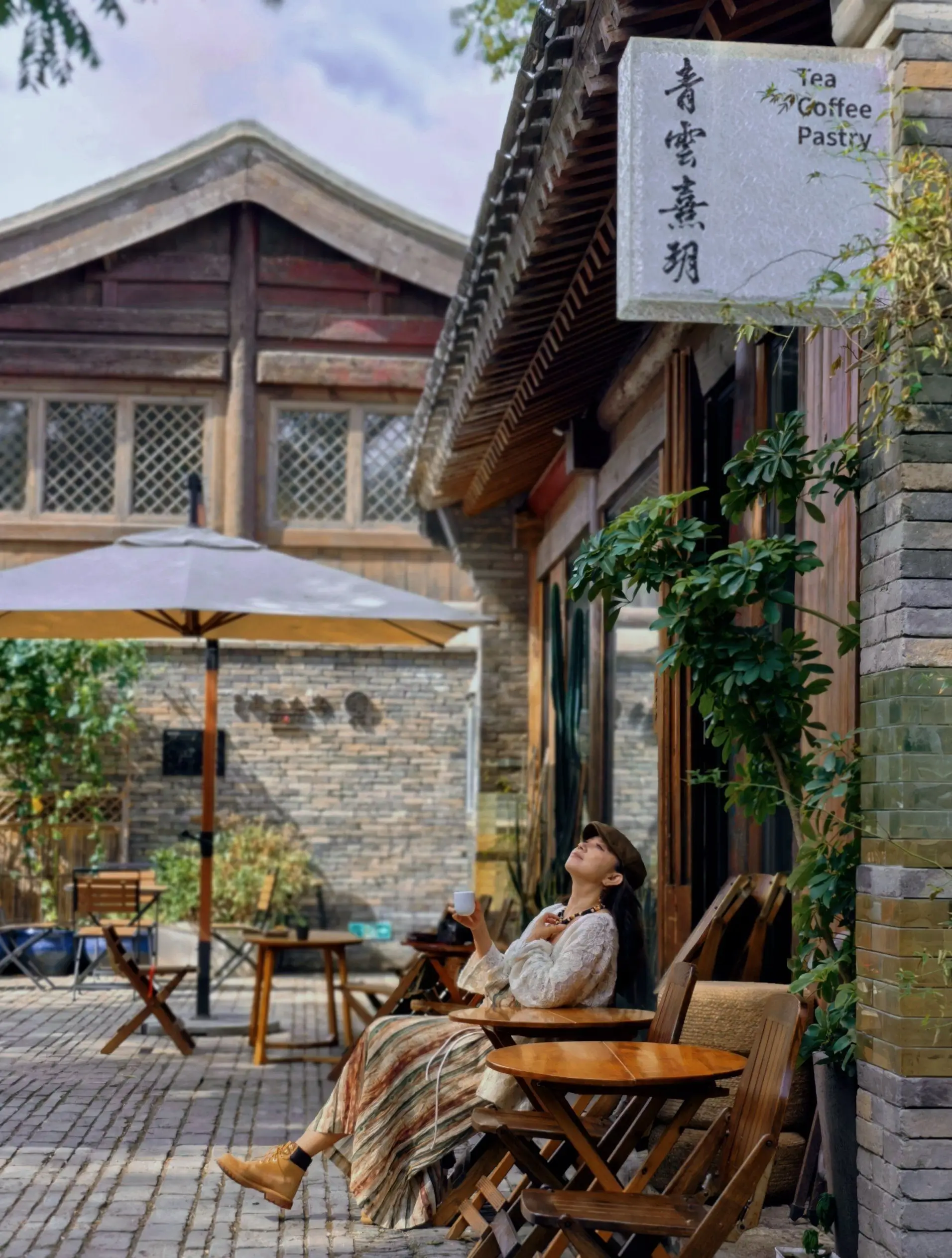 Woman relaxing with a coffee at an outdoor table in a quiet Beijing hutong courtyard café, surrounded by traditional gray brick walls and wooden buildings on a sunny day.