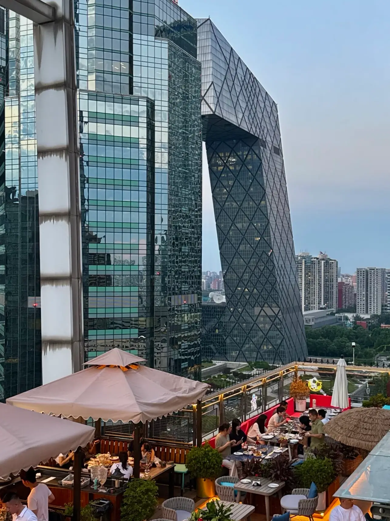 Daytime view of CCTV Headquarters (Beijing “Big Pants” building) from a rooftop terrace near China World Mall, with people dining and enjoying the Beijing skyline.