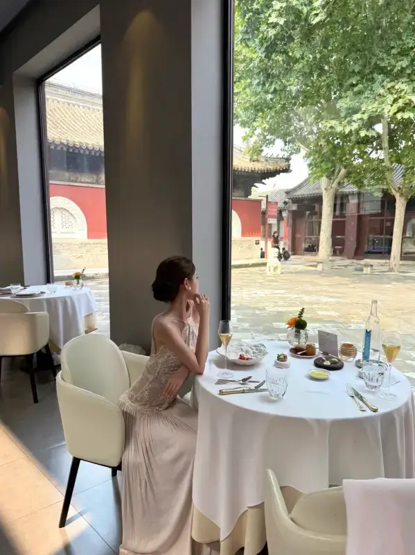 A woman seated at a window-side dining table inside Temple Dongjingyuan with a view of the courtyard