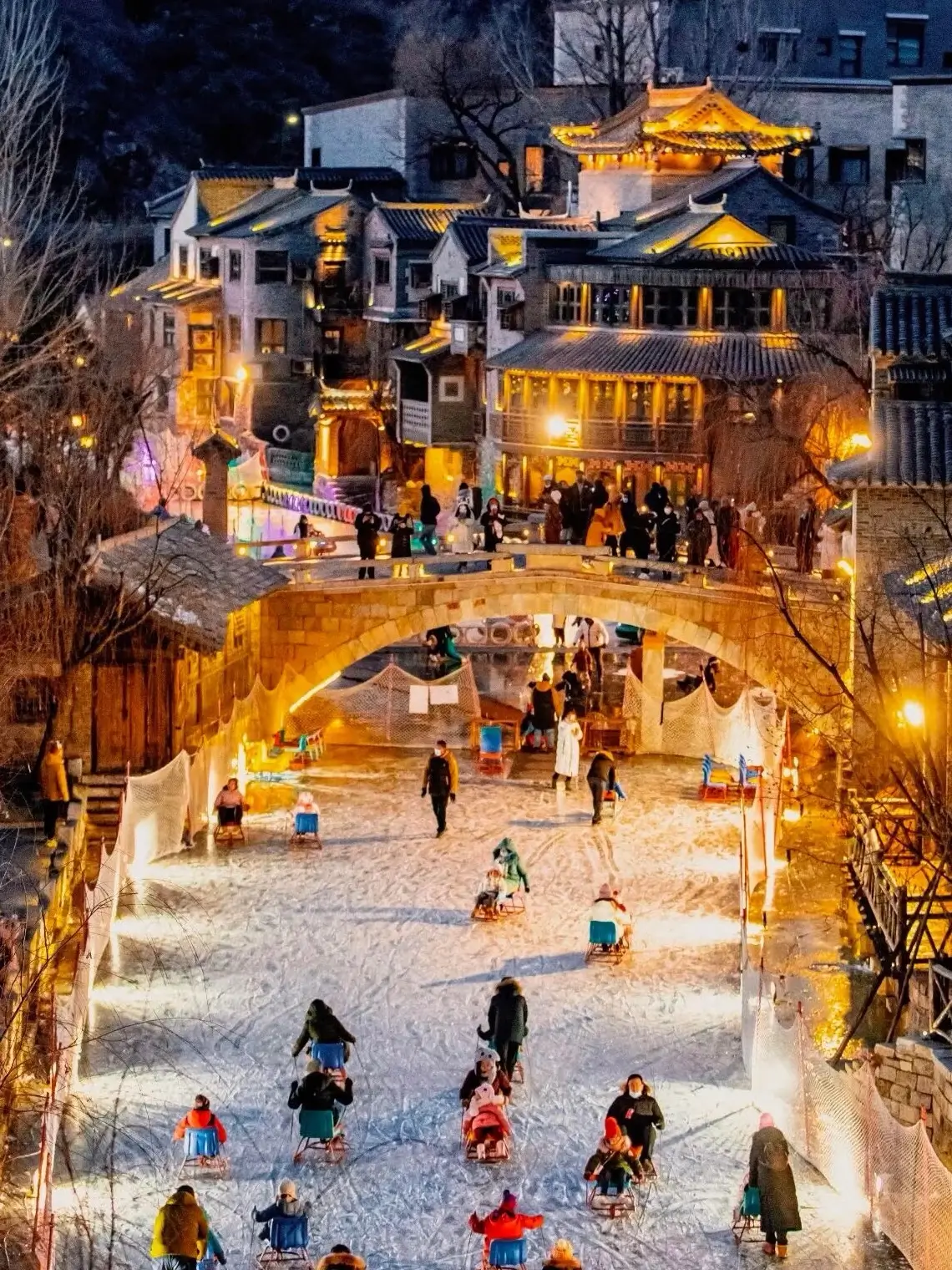 Gubei Water Town at night with a lit stone arch bridge, traditional northern-style buildings glowing with warm lights, and visitors enjoying ice and snow activities along the canal.