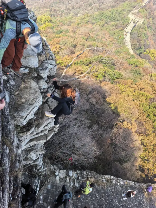 Hikers moving carefully down a rocky cliffside above forested slopes and a lower section of the Great Wall.