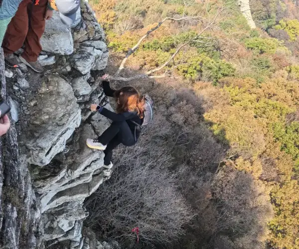Hikers moving carefully down a rocky cliffside above forested slopes and a lower section of the Great Wall.