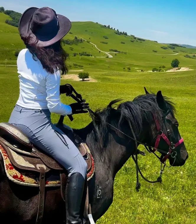 Woman horseback riding across open green grasslands near Beijing, wearing a wide-brim hat and riding a black horse along scenic countryside trails.