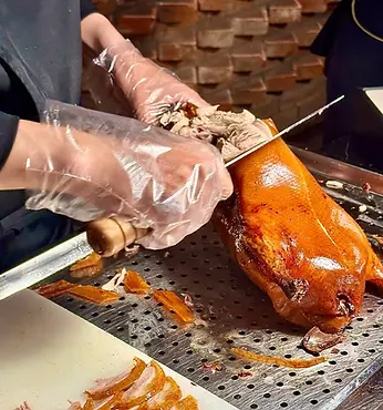 A chef wearing clear gloves skillfully slicing a crispy, golden-brown Peking duck. The text on the right side reads "Qianmen Hutong Dinner Tour" at $69/pax, featuring 4 dine-in stops and classic Beijing flavors in local eateries over 3 hours.