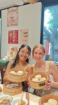 Two travelers smile while holding bamboo steamers filled with handmade baozi during a Beijing hutong bun-making experience.