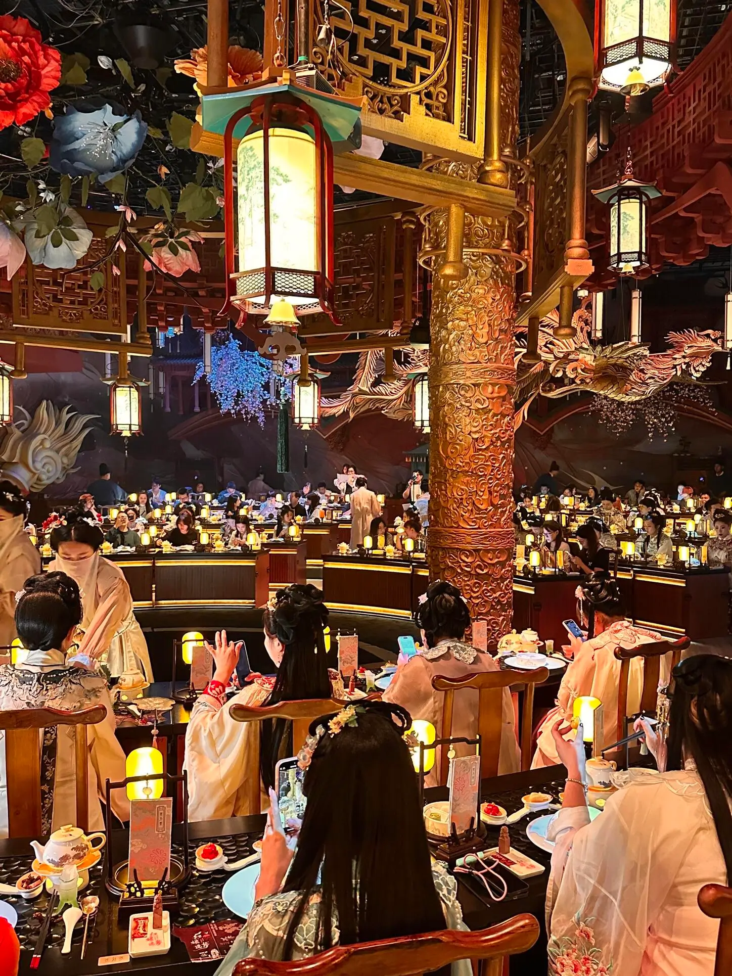 A lively dining hall featuring ornate Chinese-inspired décor, glowing lanterns, carved wooden columns, and guests enjoying a live cultural performance in a festive atmosphere.
