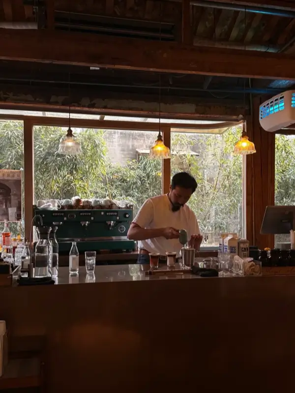 Barista preparing coffee behind a wooden counter in a sunlit hutong café in Beijing, with large windows and warm interior lighting.
