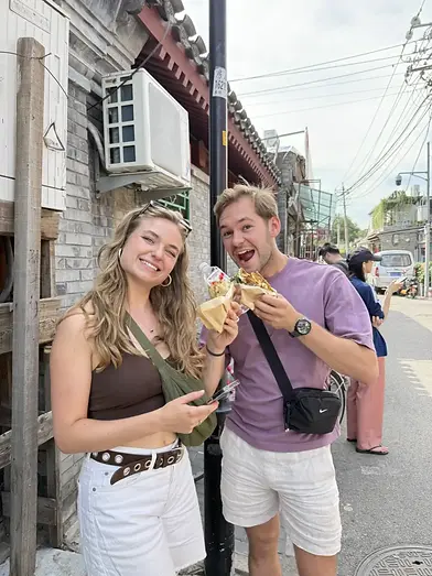 Tourists enjoying Beijing street food in a traditional hutong alley, smiling and holding jianbing