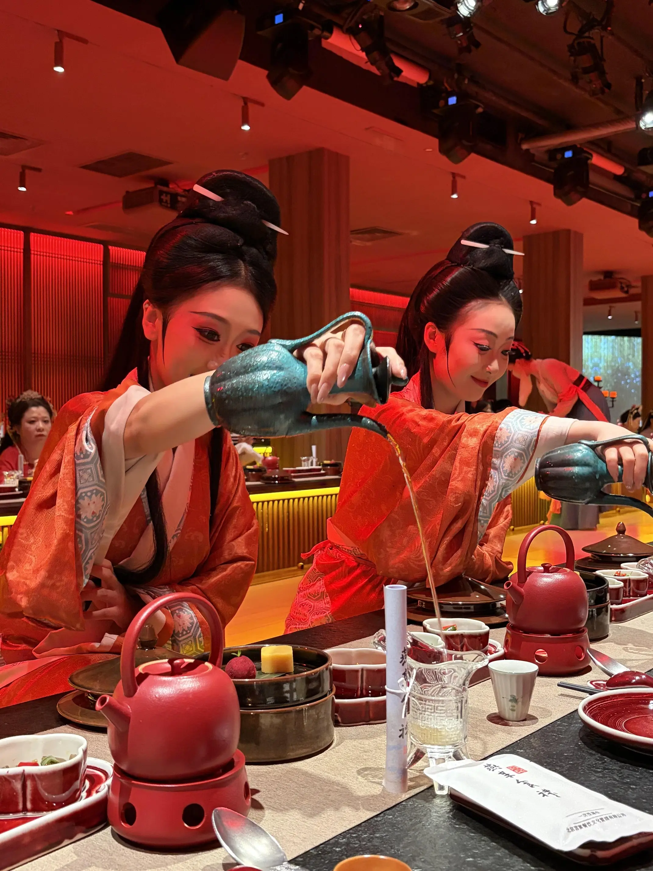 Two performers dressed in traditional Hanfu pour tea with long-spouted teapots during an interactive tea ceremony at a restaurant, with dining tables and guests in the background.