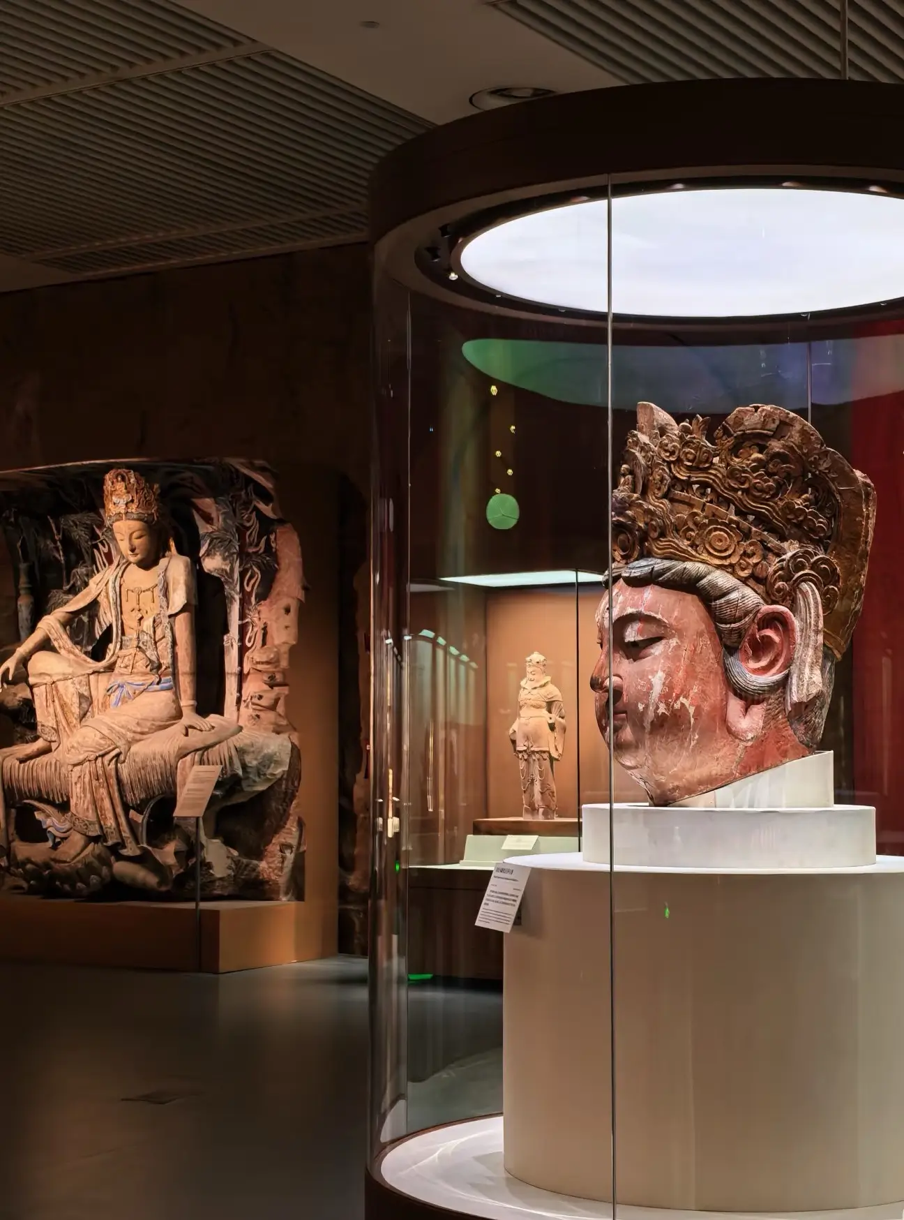 An interior view of a museum gallery showcasing ancient Chinese Buddhist sculptures. In the foreground, a large, serene head of a Buddha with a highly detailed decorative crown is displayed inside a circular glass case. In the background, a full-body stone statue of a seated Bodhisattva is positioned against a wall, illuminated by soft gallery spotlighting