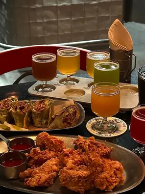 A tray of colorful craft beer samples in small glasses, served alongside crispy fried chicken, dipping sauces, and sliced wraps on a table in a modern bar setting on Lama temple food tour with Bite escape