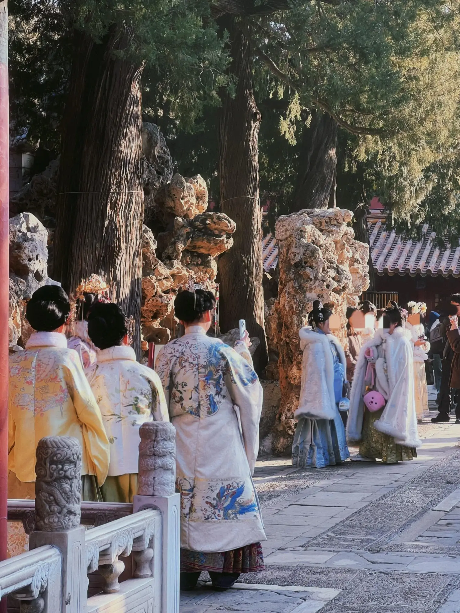 Visitors dressed in traditional Hanfu walk through a classical Chinese garden lined with tall trees, ornamental rock formations, and historic architecture.
