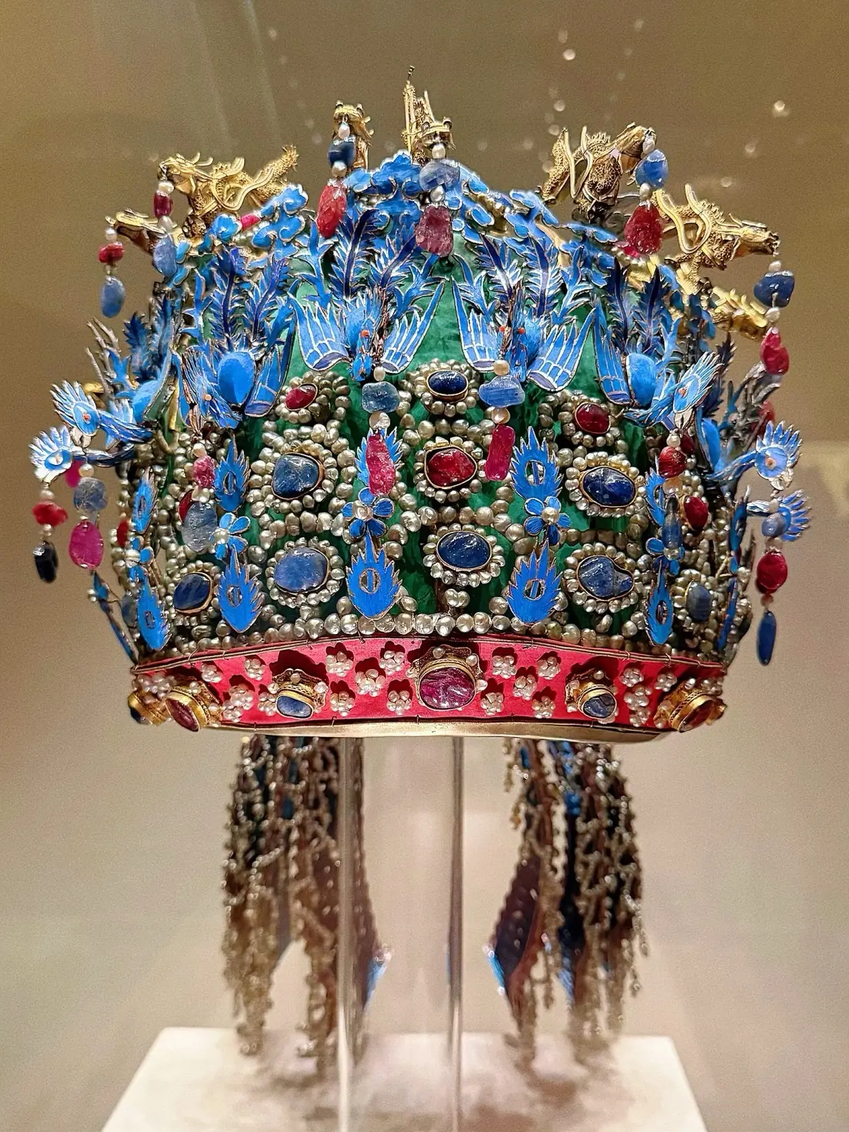A close-up of an exquisite Phoenix Crown from the Ming Dynasty, displayed on a transparent stand. The crown is ornately decorated with vibrant blue kingfisher feathers (tian-tsui), gold dragons, and inlaid with numerous red rubies, blue sapphires, and white pearls. Intricate beaded tassels hang from the sides against a neutral museum background.