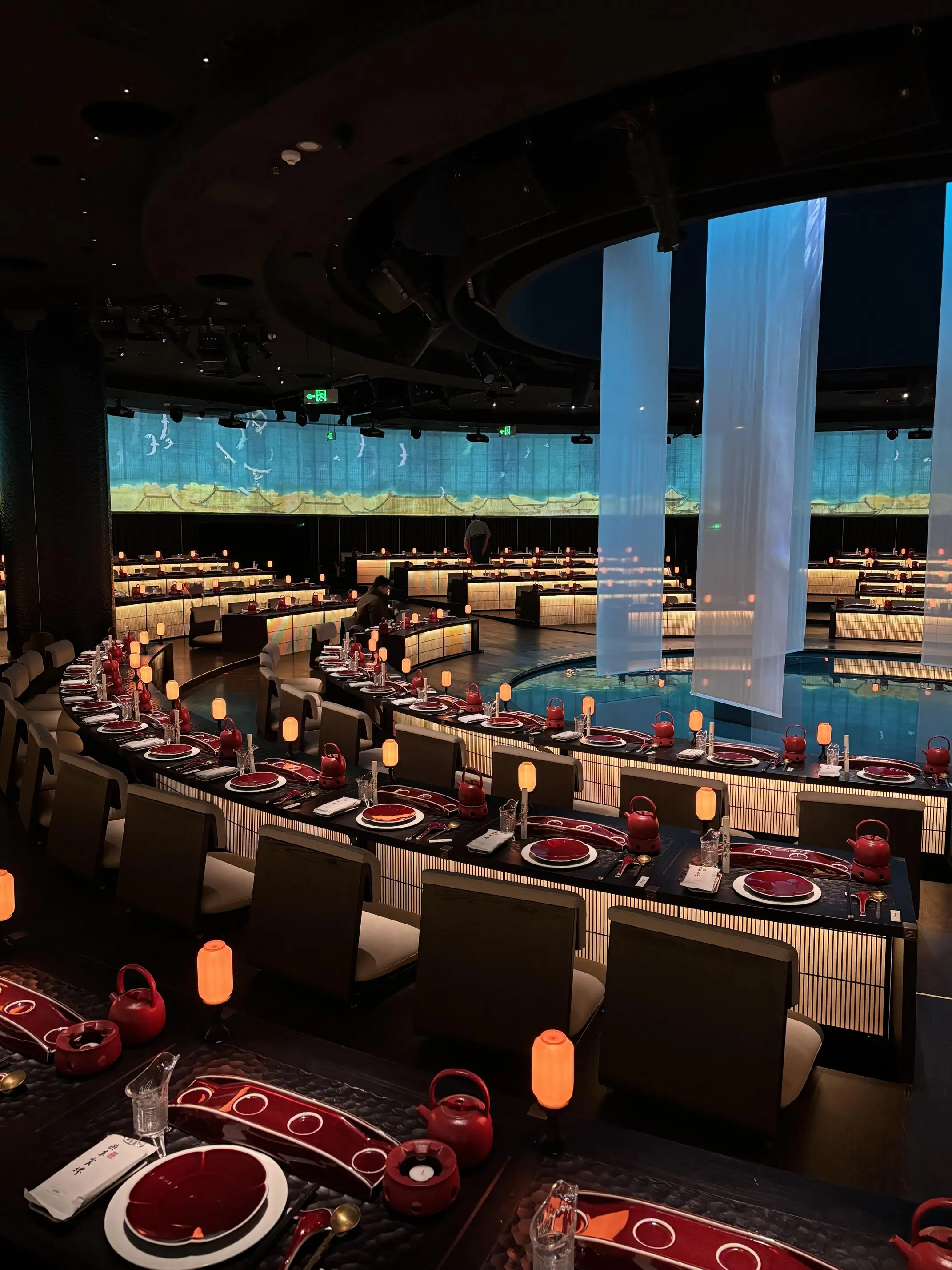 A spacious theater-style dining venue with rows of elegantly set tables surrounding a central performance area, illuminated by warm ambient lighting and large hanging fabric panels.
