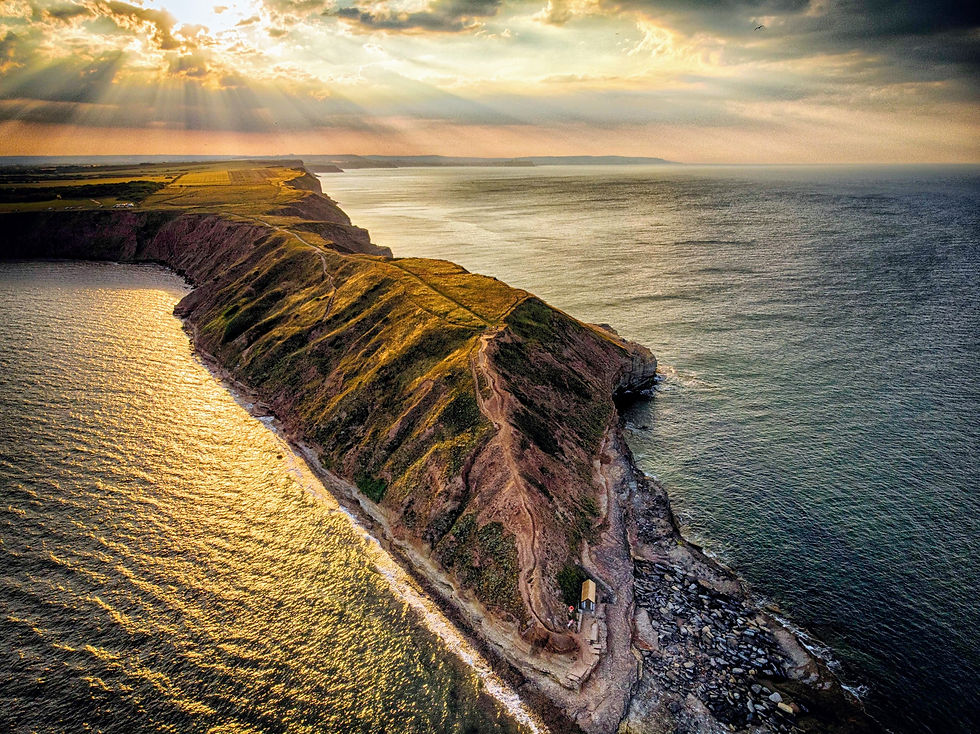 Cliffside landscape at sunset with golden light on rocky terrain, trails, and vast ocean. Dramatic sky with sun rays breaking through clouds.