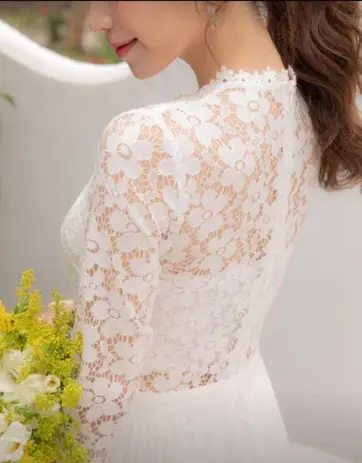 Woman wearing elegant white lace high-neck Bridal Wear, holding yellow flowers.