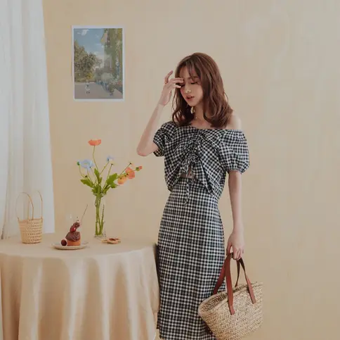 Woman in off-shoulder checkered set, woven handbag, standing in a beige room.