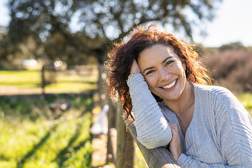 Happy curly-haired woman smiling brightly, leaning on a rustic fence.