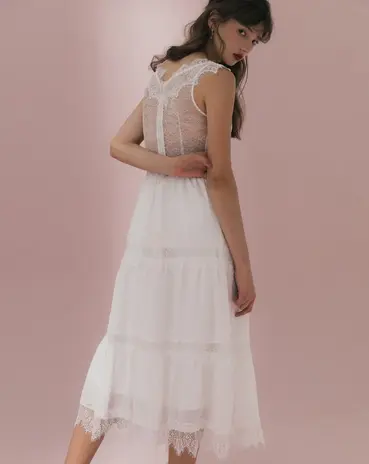 Young woman models elegant white tiered dress with sheer top, pink background.