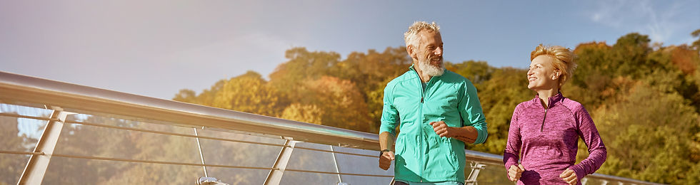 Elderly couple jogging on a sunny bridge with autumn trees in background.