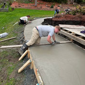 Workers leveling wet concrete sidewalk, using tools, surrounded by grass and dirt.