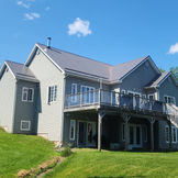 Gray house with upper deck, green sloped lawn, under blue sky.