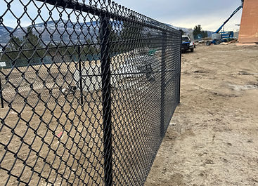 Black chain-link fence bordering a dirt construction site under cloudy sky.