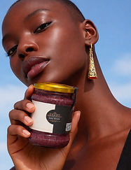 Person with dark skin holding a jar labeled “CASA MOLE” close to their face, wearing a long gold earring against a blue sky.