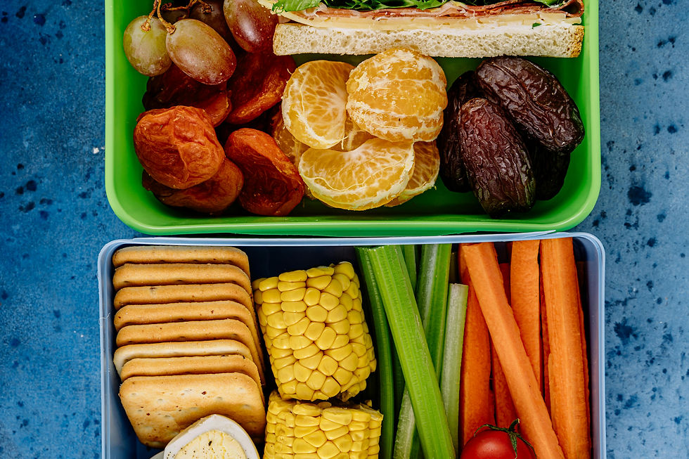 The image depicts a colorful assortment of food items. On the left side, there are peeled mandarin oranges, dried fruits, and part of a sandwich with visible lettuce and other fillings. On the right side, there are celery sticks, carrot sticks, pieces of corn on the cob, and a row of round crackers. The foods are neatly arranged in separate compartments of a container.