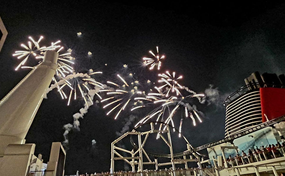 Fireworks burst over a cruise ship deck at night, with passengers watching near a roller coaster and red-striped funnel.