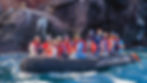 Group in red life vests on a black raft in choppy water, waving and smiling against rocky cliffs. Bright, sunny day.