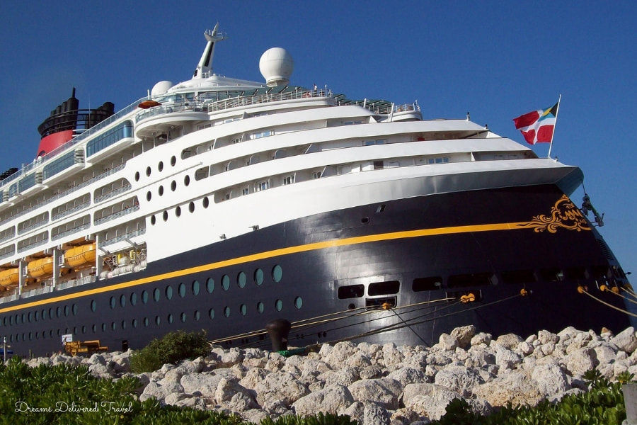 A large cruise ship docked at a port, with multiple decks and a distinctive black and white hull with gold accents. The ship has several lifeboats and is flying flags at the stern. The sky is clear and blue, and the foreground features rocky terrain and some greenery.