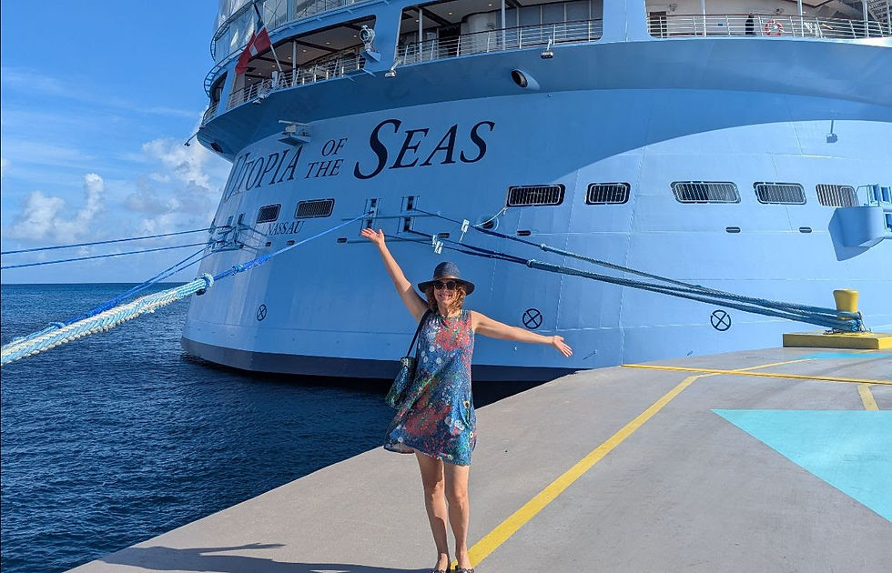 Lisa in a colorful dress and sun hat stands joyfully with arms outstretched on a dock in front of the Royal Caribbean ship, Utopia of the Seas, with a bright blue sky in the background.