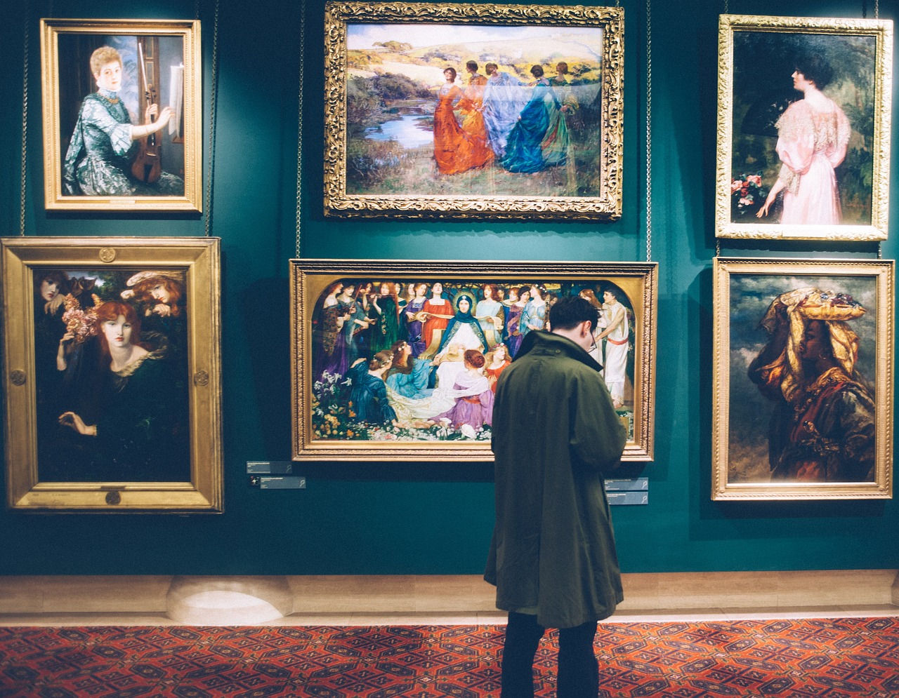 A museum visitor examining a wall of artwork. The artwork consists of various paintings in different styles and colors.