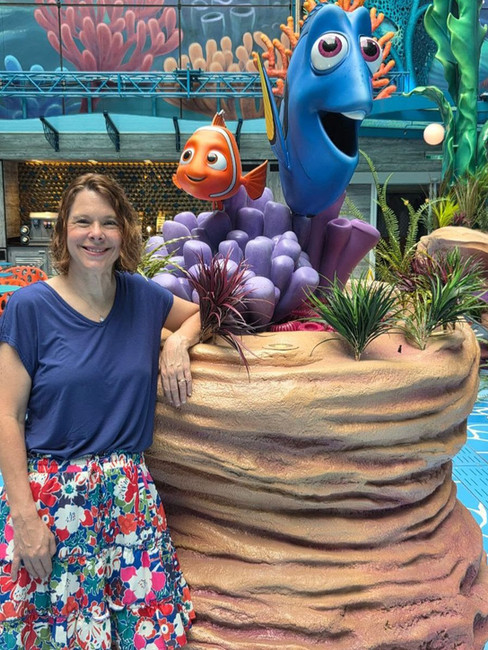 Lisa smiling beside a colorful Finding Nemo display featuring Dory and Nemo among coral and sea plants aboard a Disney-themed cruise ship. She is wearing a blue top and a floral skirt, standing next to the underwater-inspired sculpture in a bright indoor atrium.