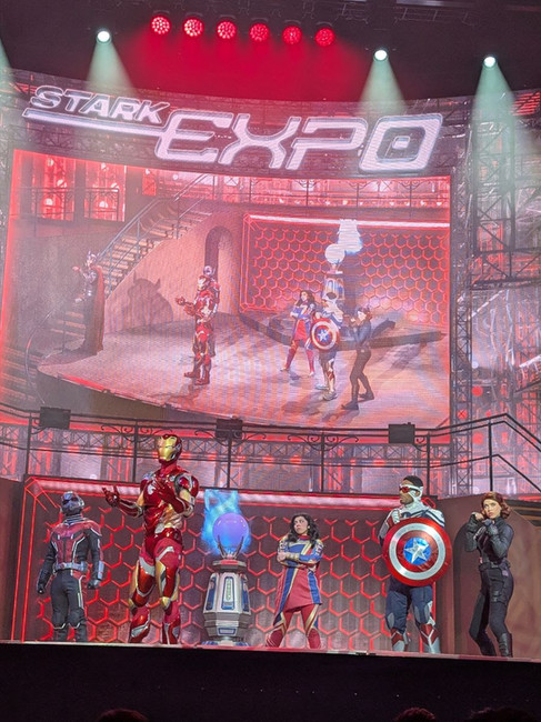 The Avengers Assemble! stage show aboard the Disney Adventure features Iron Man, Captain America, Ms. Marvel, Ant-Man, and Black Widow performing at the Stark Expo with dramatic lighting, special effects, and large video screens showcasing the action.
