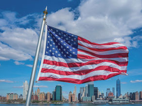 US flag waving against a blue sky with clouds. City skyline in the background. Bright colors create a sense of patriotism and pride.